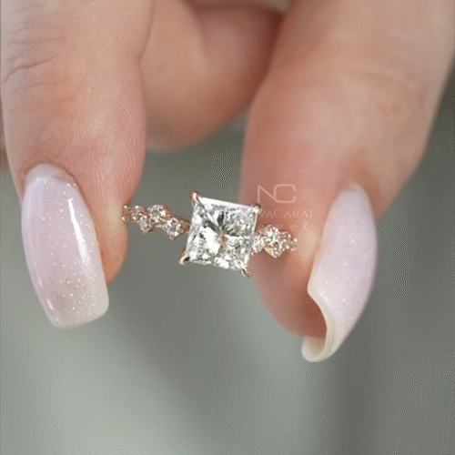 Elegant princess-cut moissanite engagement ring in rose gold, featuring a fully hidden halo for extra sparkle. The center stone is framed by a unique side profile with accent diamonds along the band, adding brilliance from every angle. A perfect blend of modern design and timeless elegance, ideal for proposals, anniversaries, or special occasions.